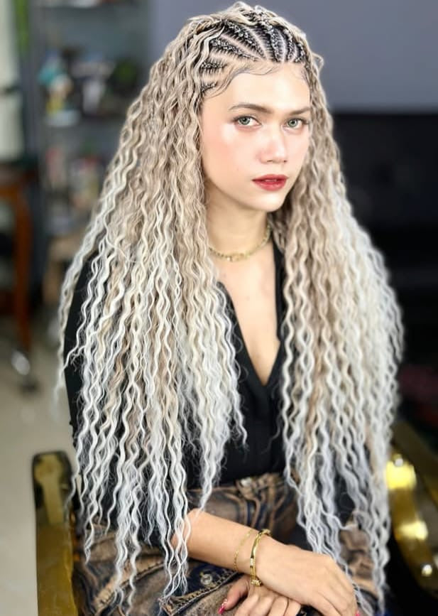 A woman with long, wavy, platinum blonde synthetic braided fulani boho braids in waist length  sits in a salon chair. The hair features dark roots and intricate cornrow braids along the top, framing her face.