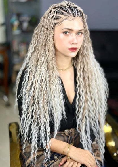 A woman with long, wavy, platinum blonde synthetic braided fulani boho braids in waist length  sits in a salon chair. The hair features dark roots and intricate cornrow braids along the top, framing her face.
