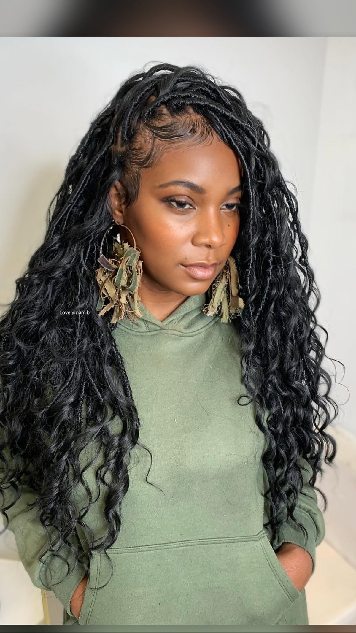 A woman with long, black, curly boho individual locs wearing a sage green hoodie and large, decorative gold hoop earrings with dried grass accents.