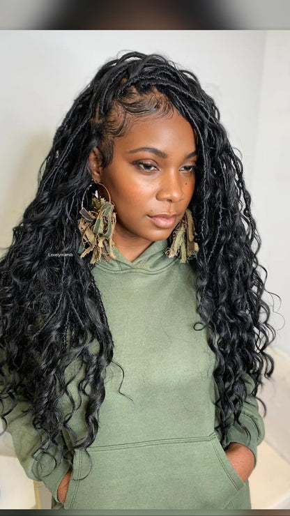 A woman with long, black, curly boho individual locs wearing a sage green hoodie and large, decorative gold hoop earrings with dried grass accents.