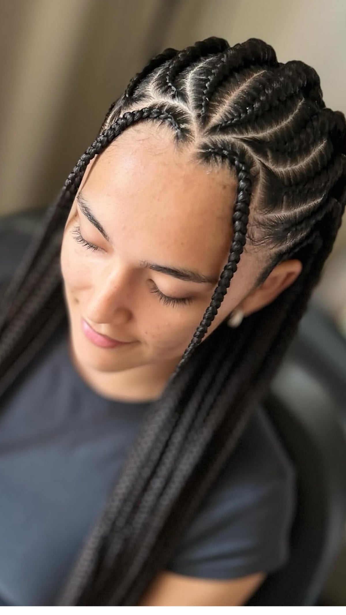 A close-up of a person with long black fulani braids or cornrow and braids, wearing a dark gray t-shirt, looking down with a calm expression.