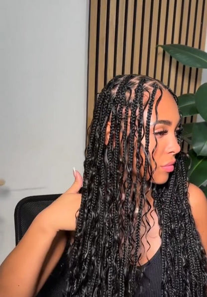 A woman with long, wet black box braids is shown in profile, holding a section of her hair. She is wearing a black top and sitting in a chair, with a slatted wood wall and a green plant in the background.
