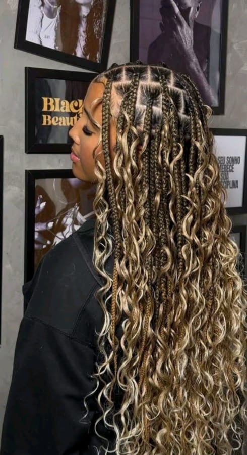 A woman shown from the side profile wearing waist length, curly boho box braids with a highlighted blonde and dark brown color scheme. She is wearing a black salon cape, with framed wall art in the background.