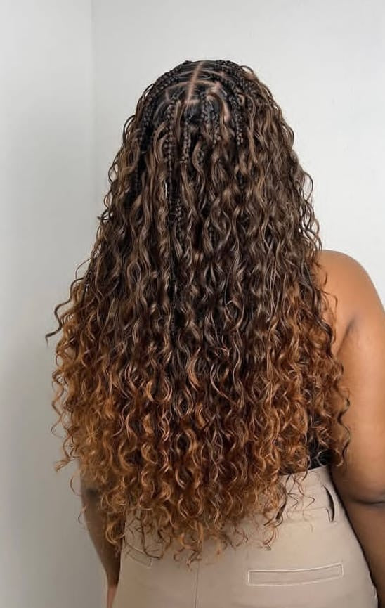 A person shown from the back wearing a long, curly boho braids in mid back length with dark brown roots and warm caramel-brown ends, paired with a beige top against a plain light grey wall.