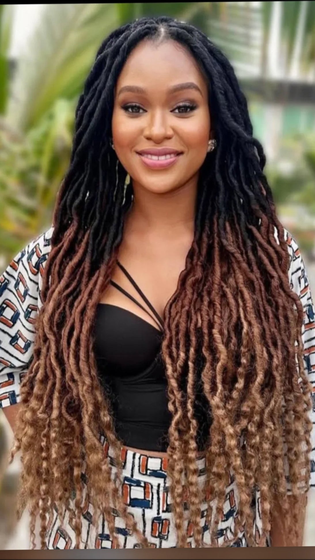 A woman with long, thick black and brown individual faux locs is shown from the chest up, wearing a black strappy top and a patterned white, black, and orange wrap jacket. The background is a soft-focus outdoor setting with green foliage.