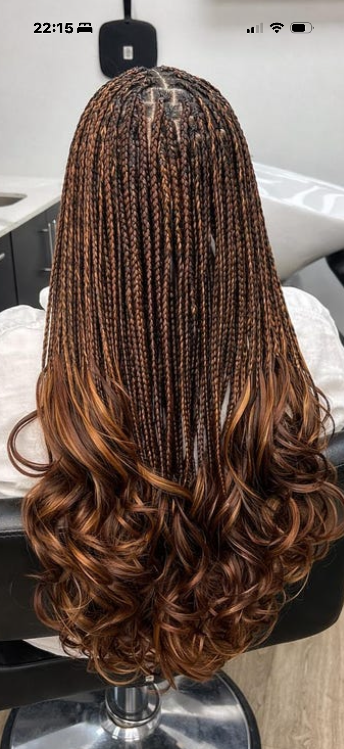 Long, wavy hair styled in tight braids, shown from the back on a salon chair. The hair features french curl waist length braids in  a dark brown root color with warm caramel and honey brown highlights throughout, transitioning into loose curls at the ends.