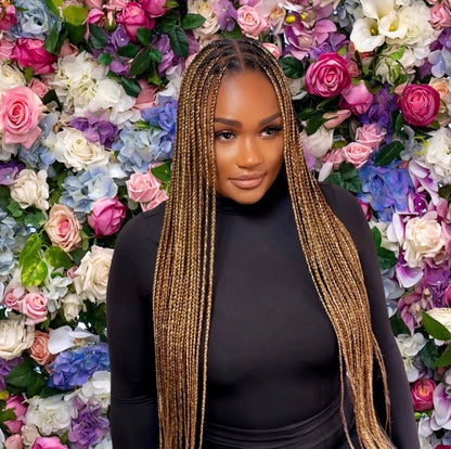 A woman with long, straight medium sized knotless  braids in a golden-brown color, wearing a black top, standing in front of a vibrant floral wall filled with pink, purple, blue, and white flowers.