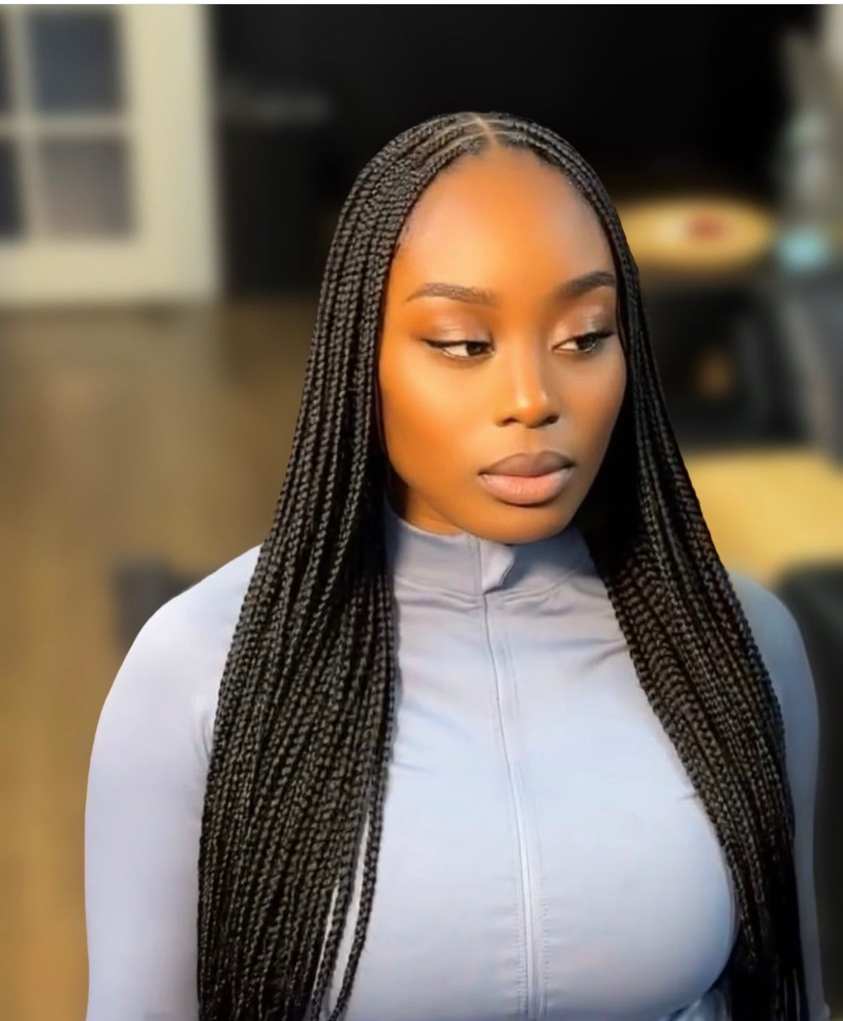 A woman with long, straight black small knotless braids wearing a light blue-gray mock-neck top, shown from the chest up against a softly blurred indoor background.