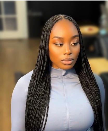 A woman with long, straight black small knotless braids wearing a light blue-gray mock-neck top, shown from the chest up against a softly blurred indoor background.