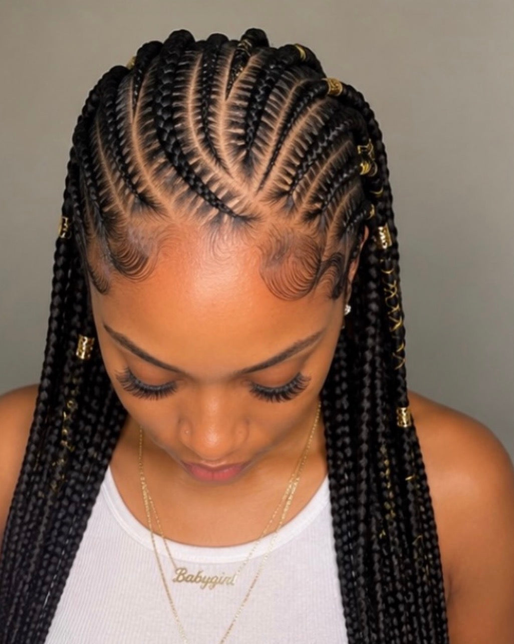 A close-up of a woman with black fulani braids styled in a cornrow pattern with small gold bead accents, wearing a white ribbed tank top and gold necklaces against a plain gray background.