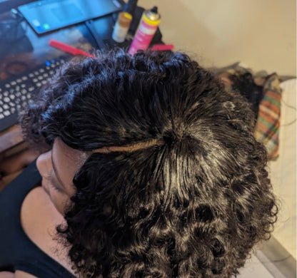 A close-up view of a person wearing a dark curly invisible part weave, showing the hair texture and part separation. The background is a cluttered indoor workspace with a keyboard, tablet, and hair styling products.