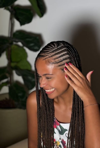 A smiling woman with long, neatly styled dark brown Fulani braids is shown indoors, touching her hair. She is wearing a white floral top, and a large green potted plant is visible in the softly lit background.