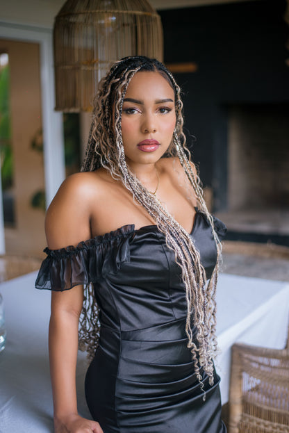 A woman with long blonde and black boho box braids wearing a black off-the-shoulder satin dress, standing in a modern indoor setting with a wicker pendant light and table visible.