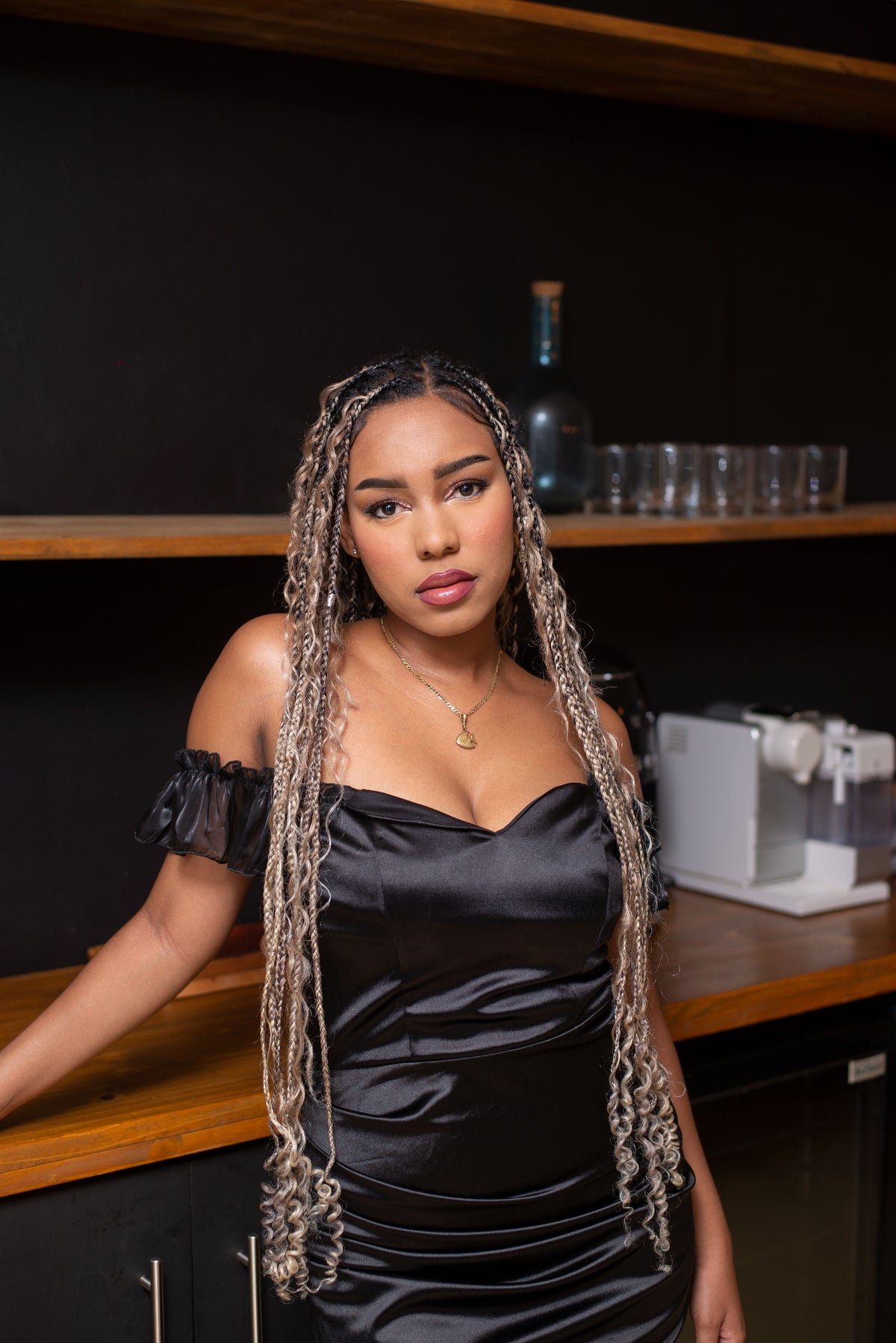 A woman with long, curly blonde and dark brown goddess braids wears a black satin off-the-shoulder dress, standing in front of a dark bar counter with wooden shelves in the background.