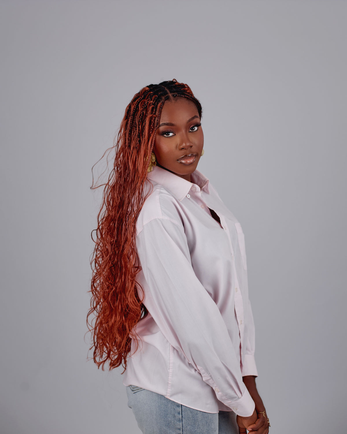 A woman with long, curly knotless boho braids in red hair with black roots is wearing a light pink button-up shirt and light blue jeans, photographed against a solid grey studio background.