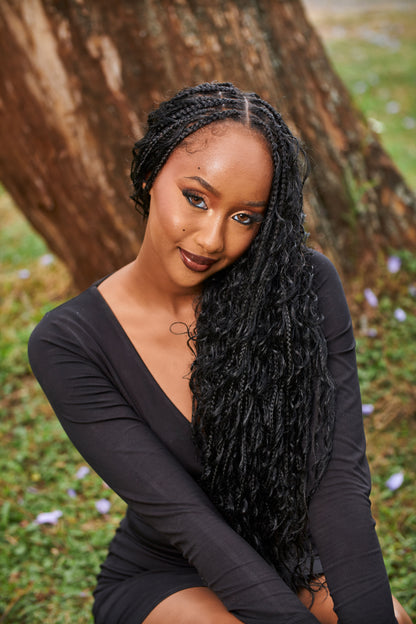 A woman with long, curly black goddess box braids sits outdoors in front of a large tree trunk, wearing a black long-sleeve dress.